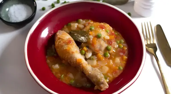 Un delicioso pollo a la jardinera en un cuenco rojo, con guisantes, zanahoria y laurel.