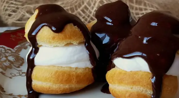 Deliciosos profiteroles de nata cubiertos con chocolate negro derretido, servidos en un plato blanco con detalles dorados.