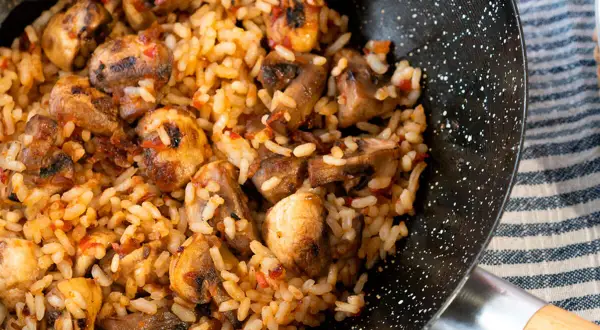Arroz con champiñones recién hecho en sartén, con trozos dorados de champiñones y un toque de pimiento rojo.