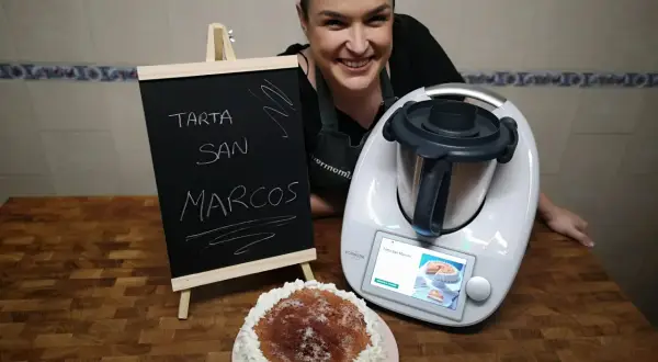Mujer sonriente junto a una tarta San Marcos y una Thermomix. La pizarra anuncia "Tarta San Marcos".