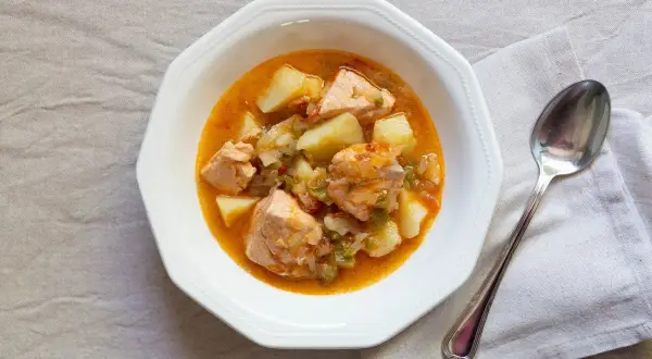 Un plato de marmitako de salmón con patatas y pimientos en una cazuela blanca, acompañado de una cuchara.