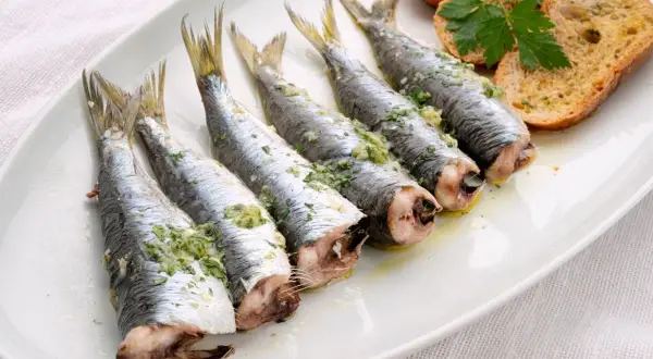Cinco sardinas al horno, preparadas con hierbas y ajo, servidas en una bandeja blanca con tostadas.