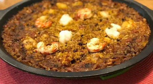 Arroz de tamboril con gambas y trozos blancos de pescado, servido en una paellera negra sobre una alfombrilla roja.