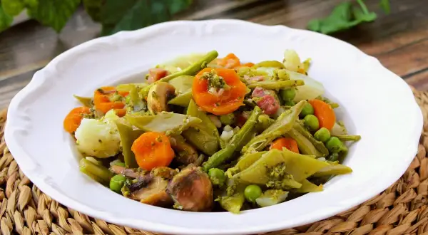 Un plato de menestra de verduras con patatas, zanahorias, judías verdes, guisantes y champiñones, todo aderezado con una salsa verde.