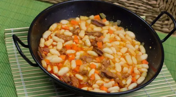Un plato humeante de alubias con chorizo, zanahoria y patata en una paellera negra, servido sobre una esterilla verde.