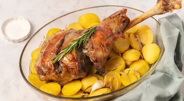 Pierna de cordero al horno dorada y jugosa, servida con patatas asadas, romero y ajo en una fuente de cristal.