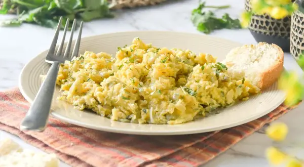 Un plato de revuelto de bacalao con perejil picado, acompañado de una rebanada de pan y un tenedor.