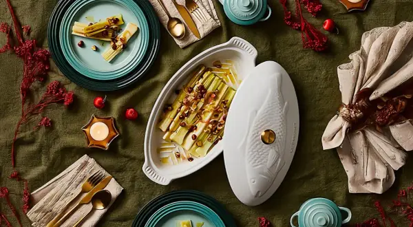 Puerros al horno, tiernos y dorados, servidos en una elegante fuente con tapa en forma de pez. Decoración festiva con velas y frutos rojos.