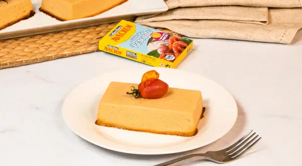 Pastel de merluza servido en plato blanco, decorado con tomate y hierbas. Caja de caldo de pescado Avecrem al fondo.