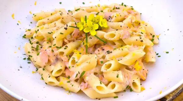 Pasta penne cremosa con trozos de salmón rosado, ralladura de limón y flores amarillas. Un plato delicioso.