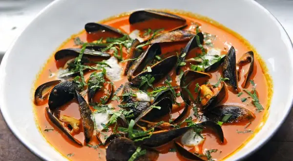 Un tazón blanco rebosante de mejillones al vapor en una salsa roja vibrante, adornado con hierbas frescas picadas.