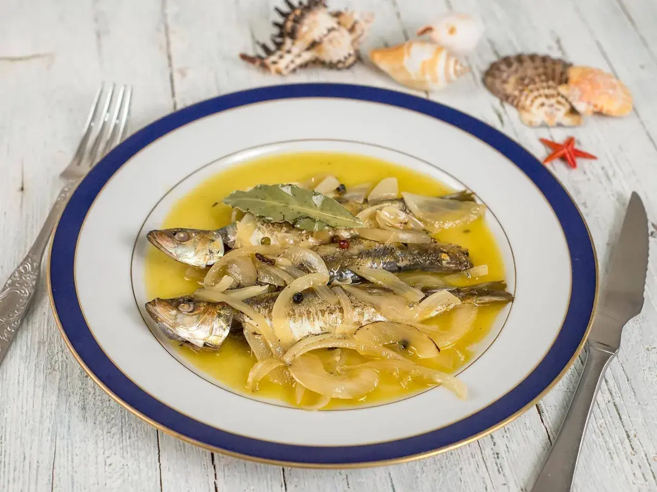 Tres sardinas en escabeche, cubiertas de cebolla y laurel, nadan en un líquido dorado. El plato está decorado con conchas marinas.