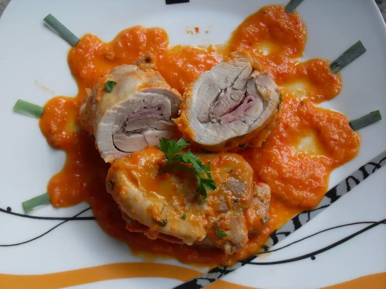 Contramuslos de pollo Thermomix enrollados con jamón, cubiertos de salsa de pimientos y decorados con perejil y cebolleta.