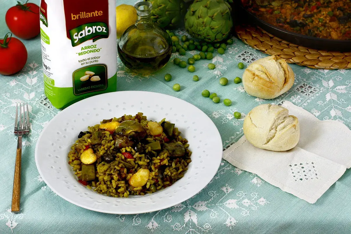 Un plato de paella de verduras, con arroz Sabroz, tomates, alcachofas, guisantes y panecillos.