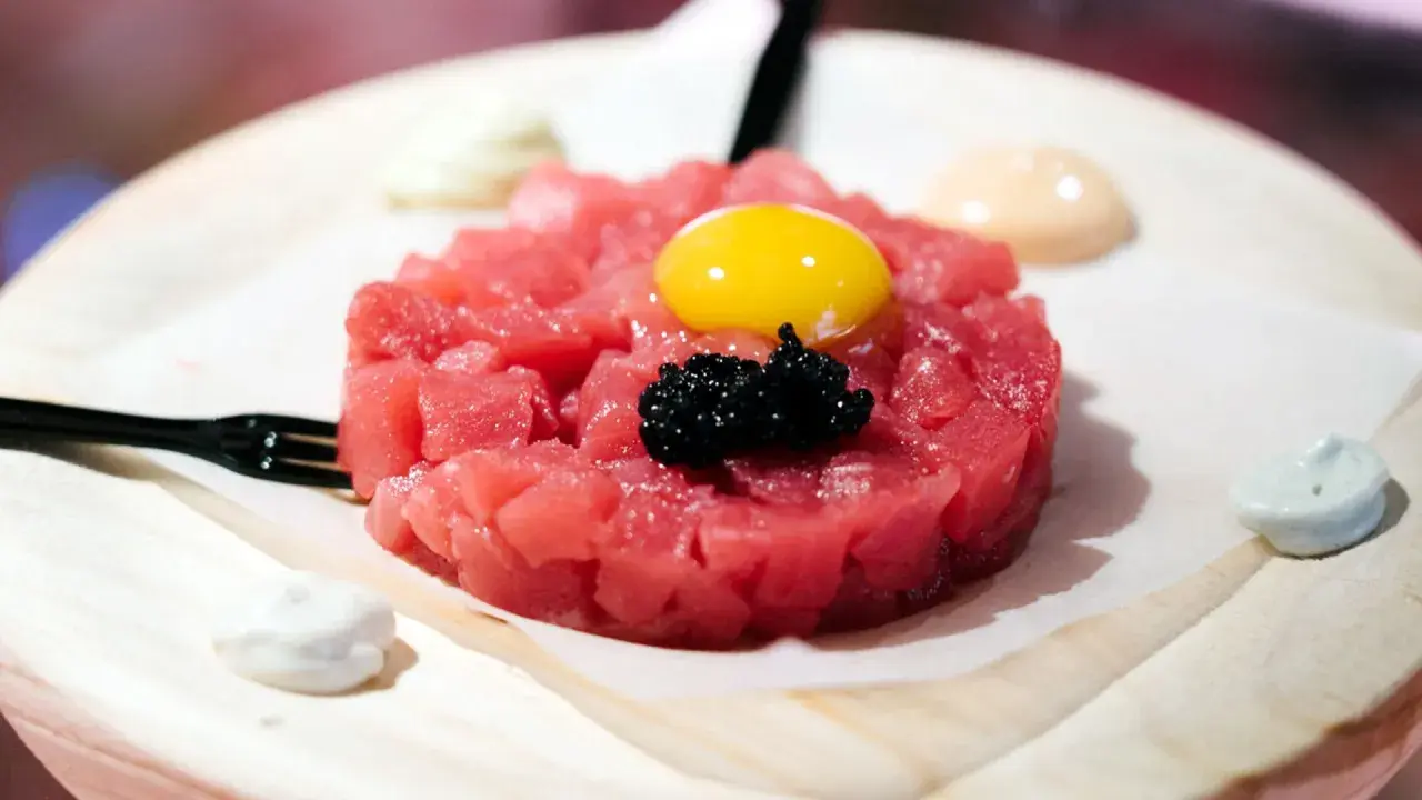 Delicioso tartar de atún fresco, coronado con yema de huevo y caviar negro. Servido con salsas cremosas y un tenedor.
