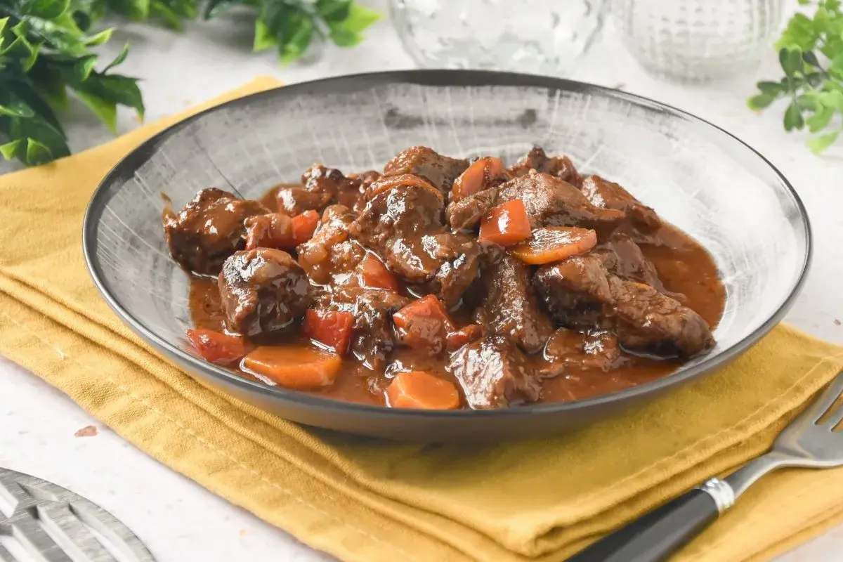 Plato humeante de carne al toro, tiernos trozos de carne en salsa oscura con zanahorias, servido en un cuenco gris sobre servilleta amarilla.