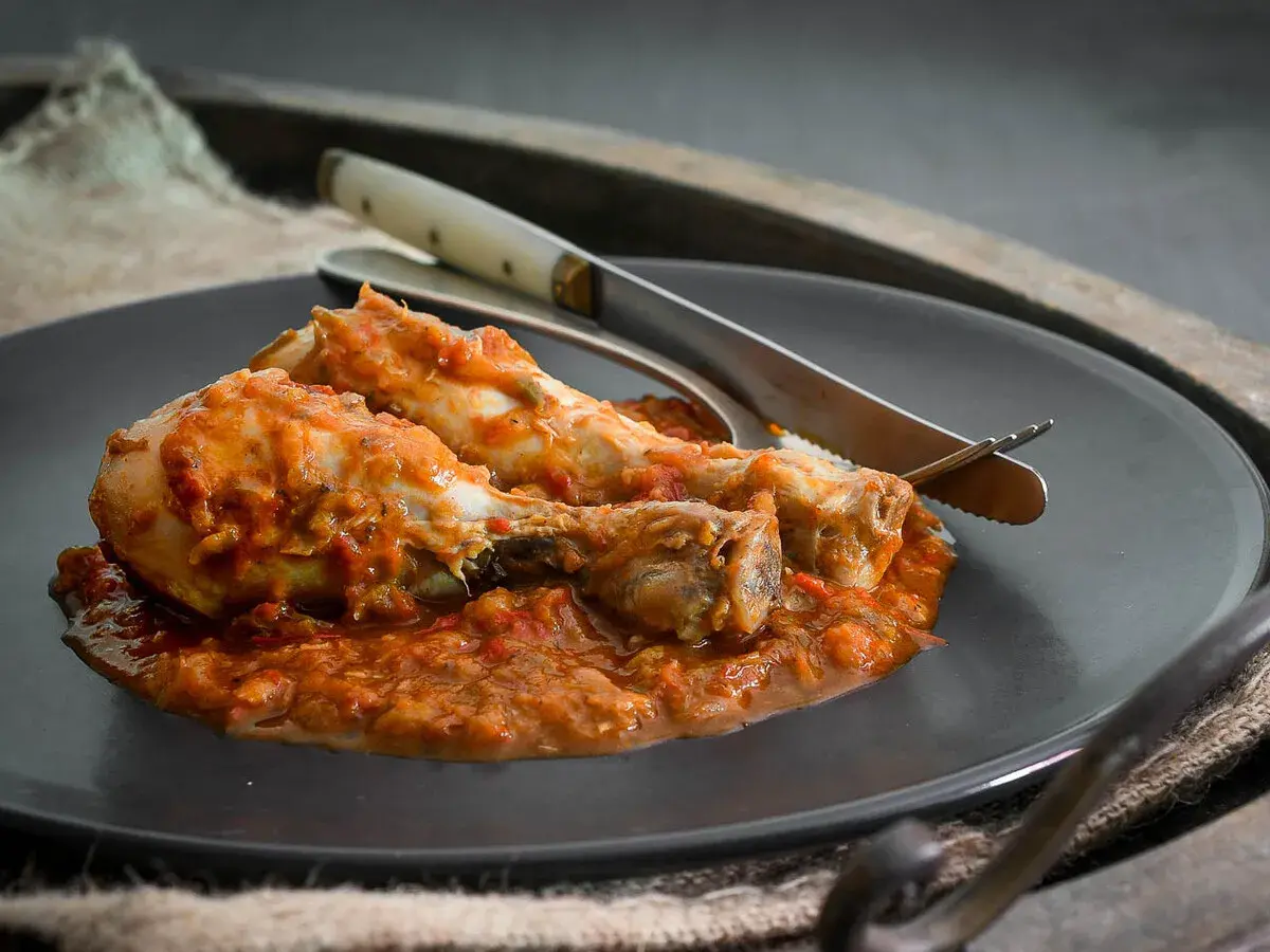 Dos muslos de pollo al chilindrón, servidos en un plato oscuro con cubiertos.