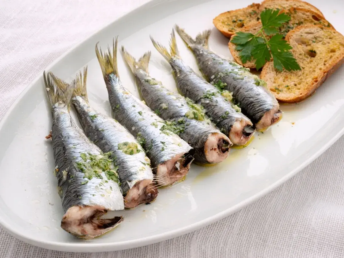 Cinco sardinas al horno, preparadas con hierbas y ajo, servidas en una bandeja blanca con tostadas.