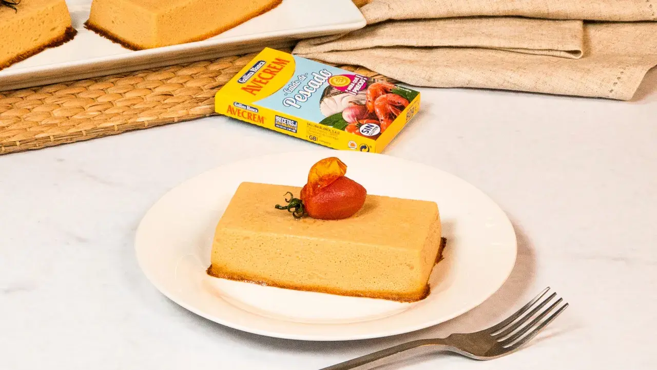 Pastel de merluza servido en plato blanco, decorado con tomate y hierbas. Caja de caldo de pescado Avecrem al fondo.