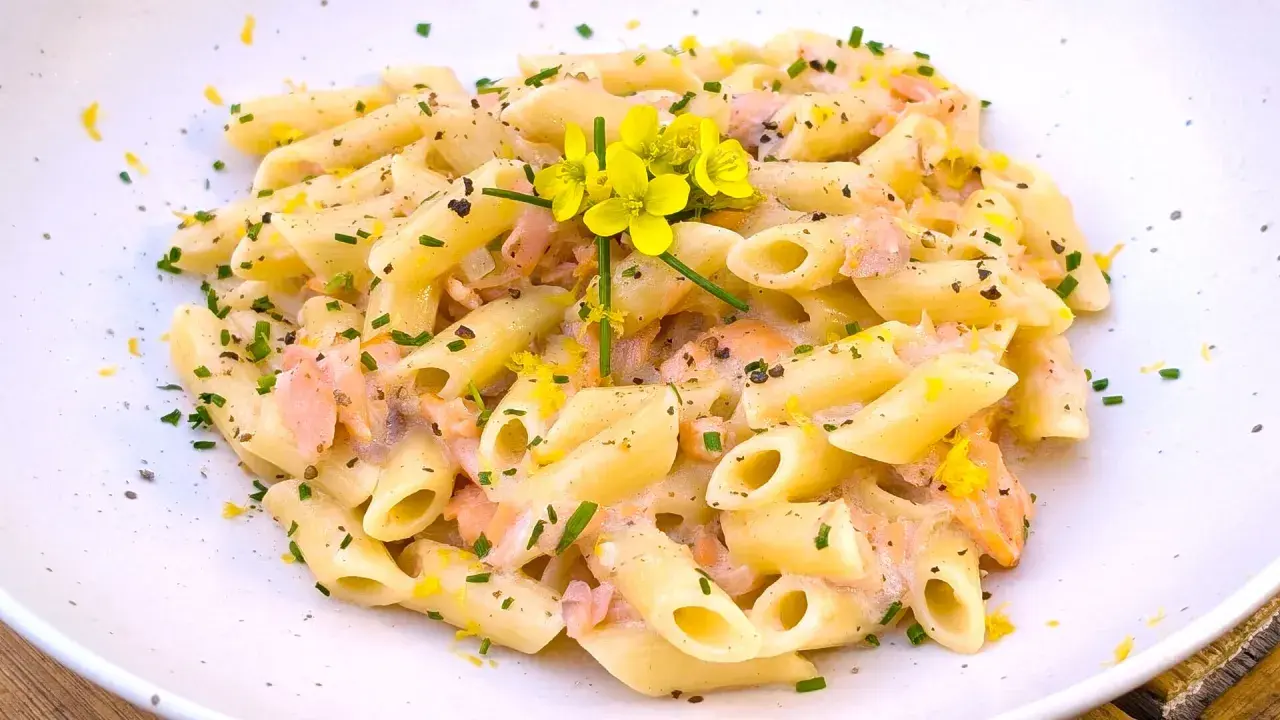Pasta penne cremosa con trozos de salmón rosado, ralladura de limón y flores amarillas. Un plato delicioso.