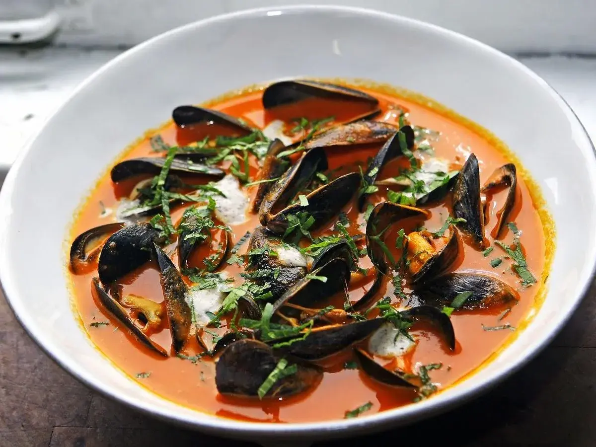Un tazón blanco rebosante de mejillones al vapor en una salsa roja vibrante, adornado con hierbas frescas picadas.