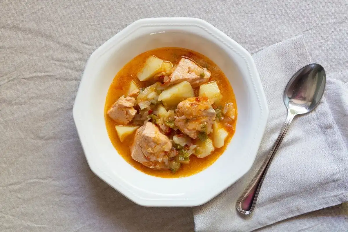 Un plato de marmitako de salmón con patatas y pimientos en una cazuela blanca, acompañado de una cuchara.