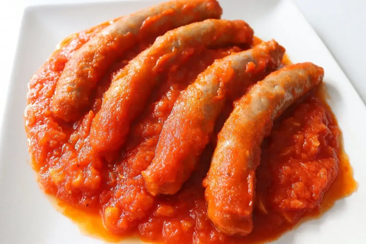 Cuatro salchichas ba&ntilde;adas en una salsa de tomate casera, servidas en un plato blanco.