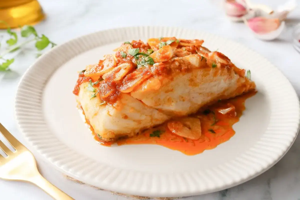 Bacall&agrave; a la llauna servido en plato blanco con salsa de tomate y ajo. Un tenedor dorado a la izquierda.