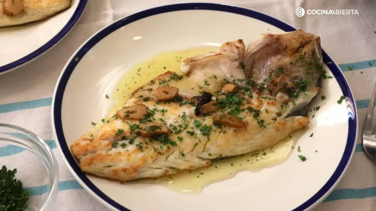 Deliciosa merluza a la plancha con ajo laminado y perejil fresco, servida en un plato blanco con borde azul.