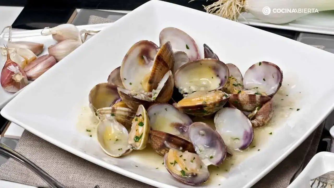Deliciosas almejas a la marinera, abiertas y jugosas, servidas en una fuente blanca con perejil picado.