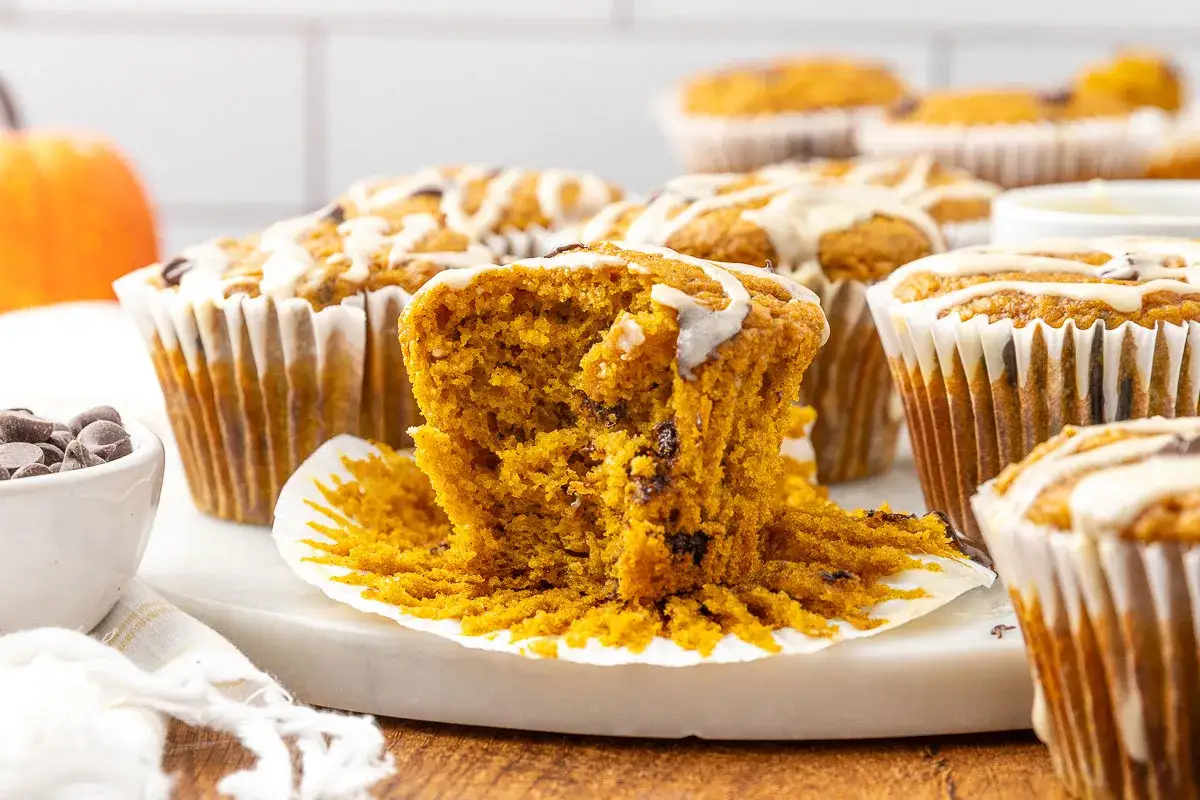Magdalenas de calabaza saludables con glaseado blanco y chispas de chocolate. Un muffin est&aacute; partido, mostrando su interior esponjoso.
