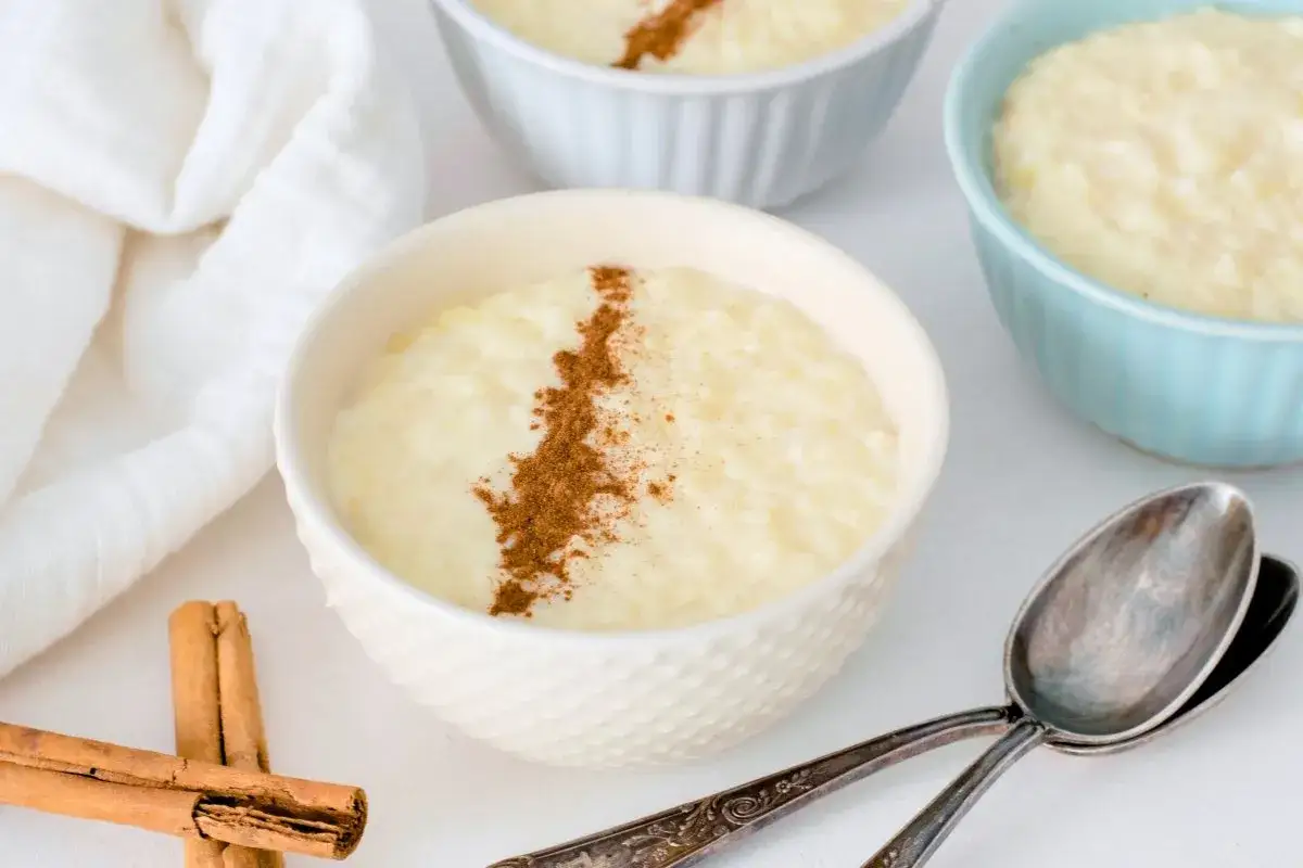 Delicioso arroz con leche cremoso, preparado en Thermomix y espolvoreado con canela. Servido en cuencos blancos y azules, con canela en rama y cucharas antiguas.