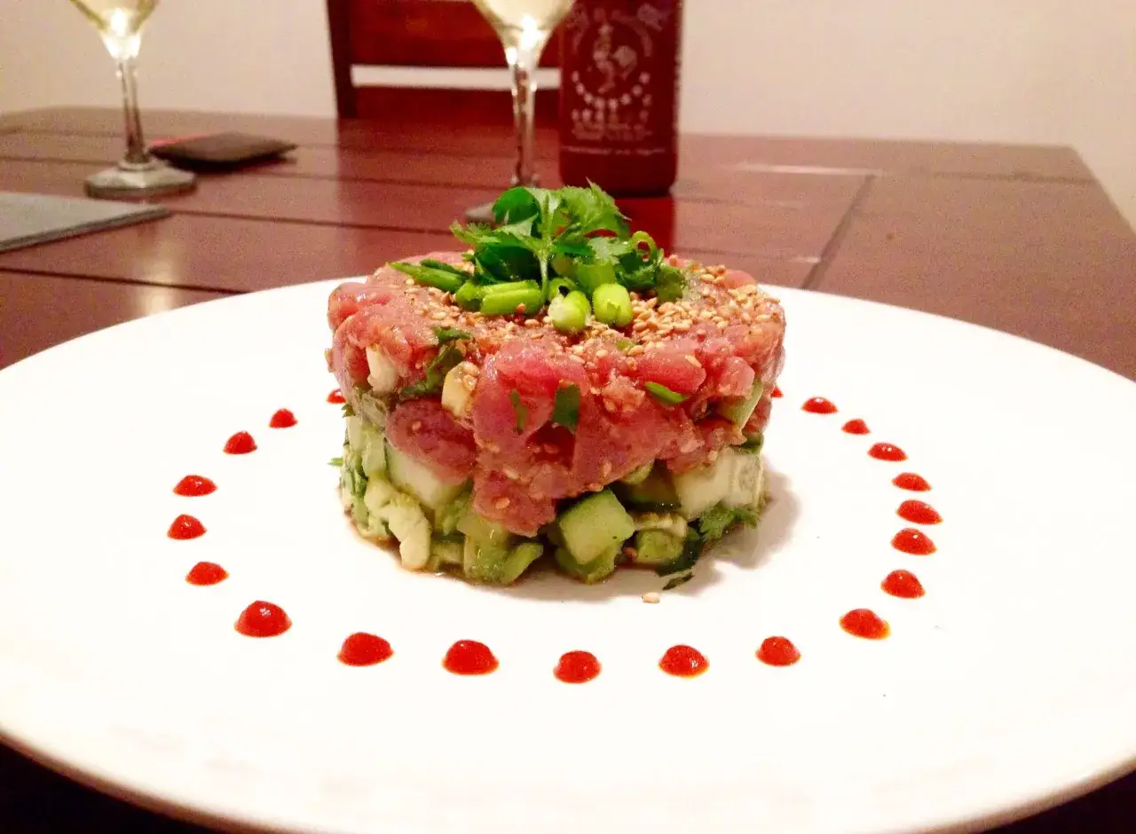 Delicioso tartar de at&uacute;n fresco con aguacate y pepino, decorado con s&eacute;samo y cilantro. Gotas de salsa picante bordean el plato.