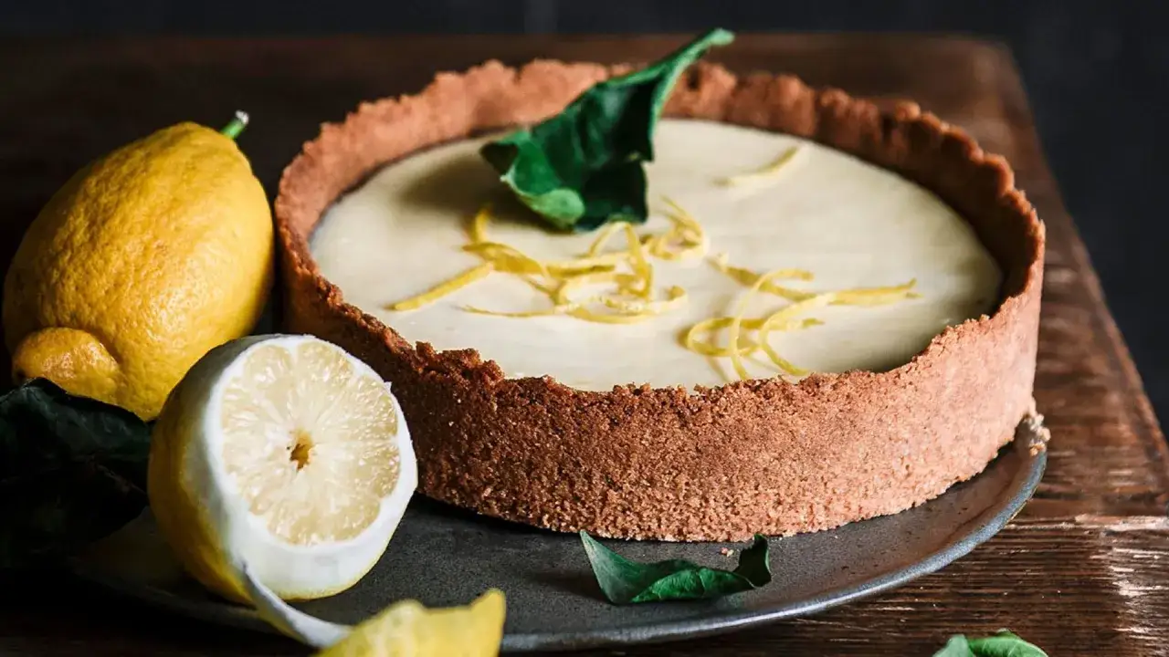 Tarta de limón cremosa con base crujiente, decorada con ralladura y hojas verdes. Limones frescos acompañan este postre.