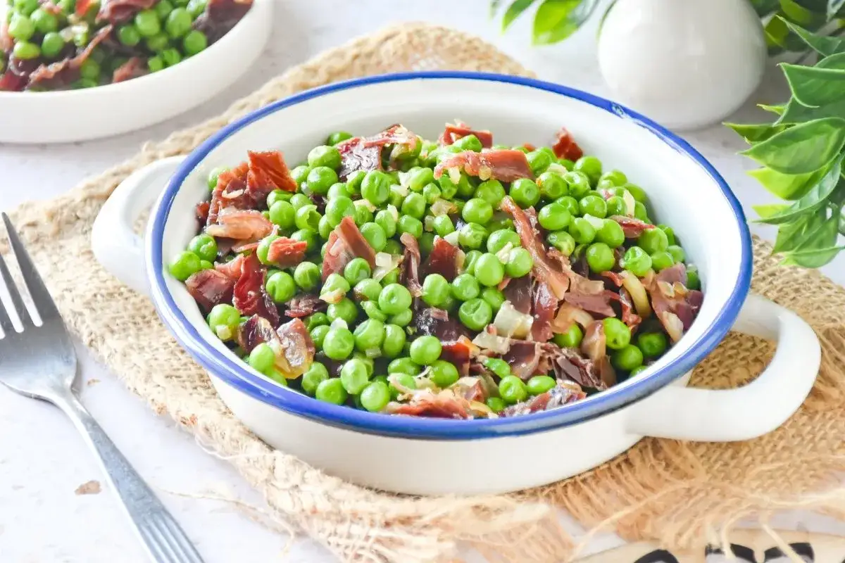 Un plato de guisantes con jam&oacute;n, un cl&aacute;sico espa&ntilde;ol, servido en un cuenco blanco con borde azul.