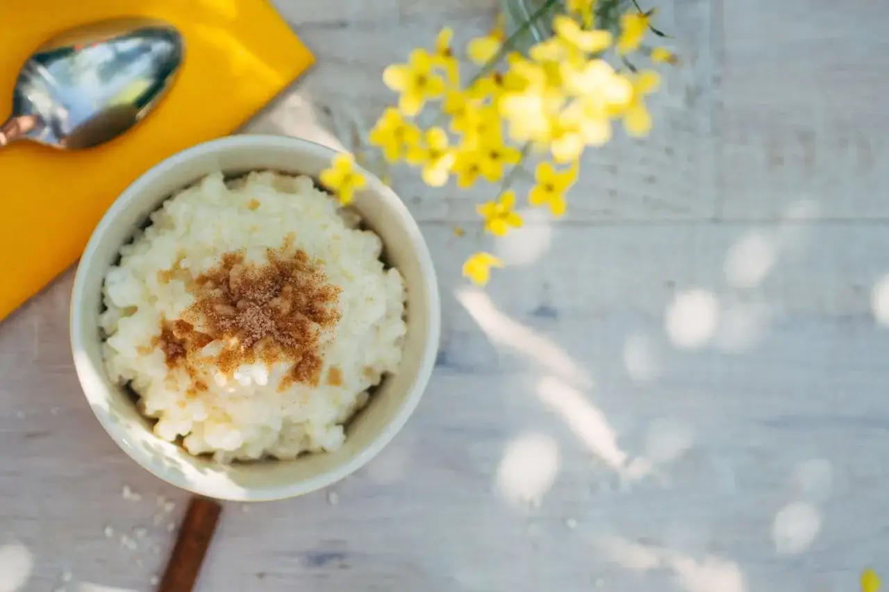 Un taz&oacute;n de arroz con leche de soja, espolvoreado con canela, junto a una cuchara y flores amarillas.