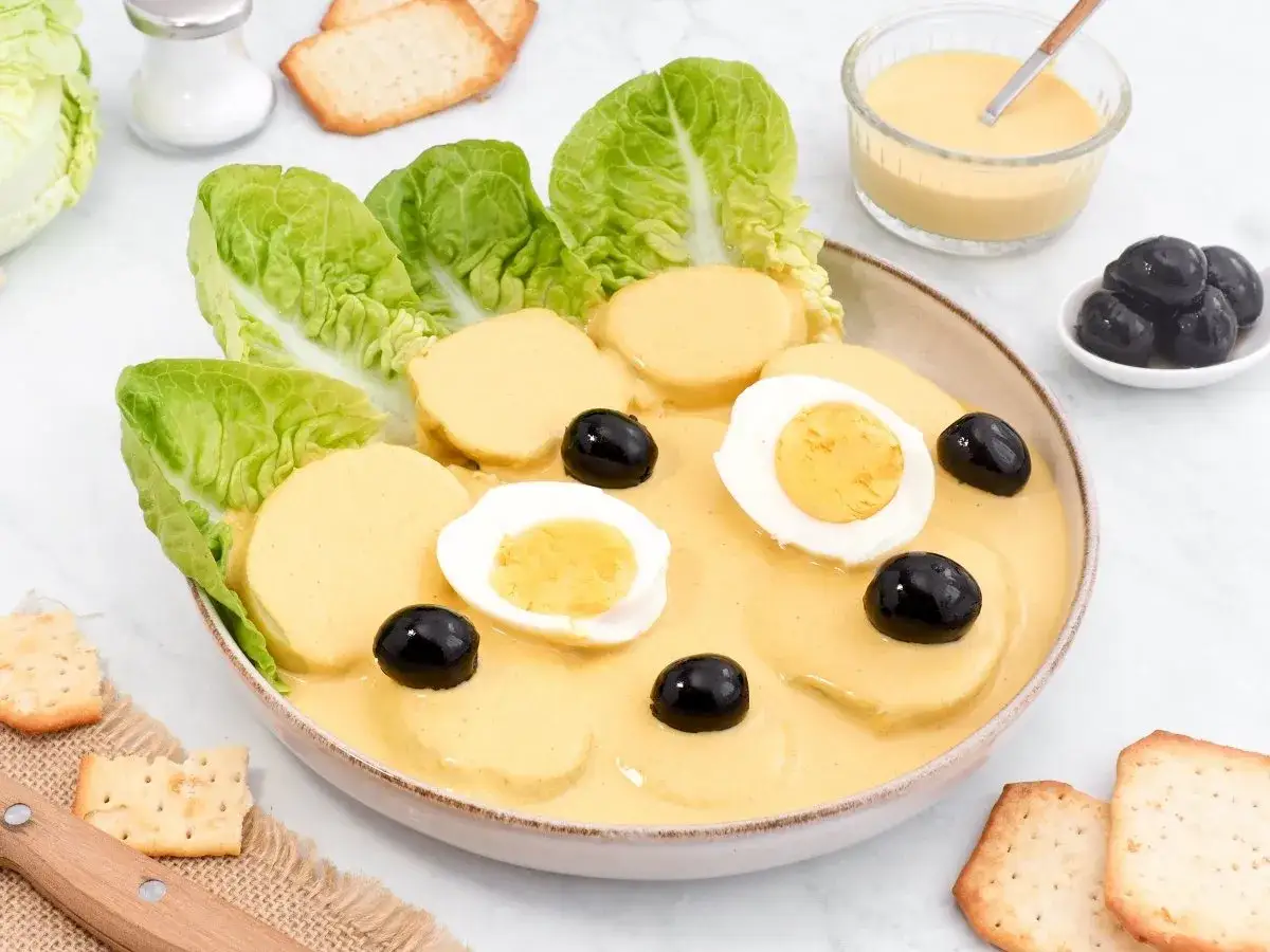 Papa a la huancaína: rodajas de papa cubiertas con salsa cremosa, adornadas con huevos duros, aceitunas negras y hojas de lechuga.
