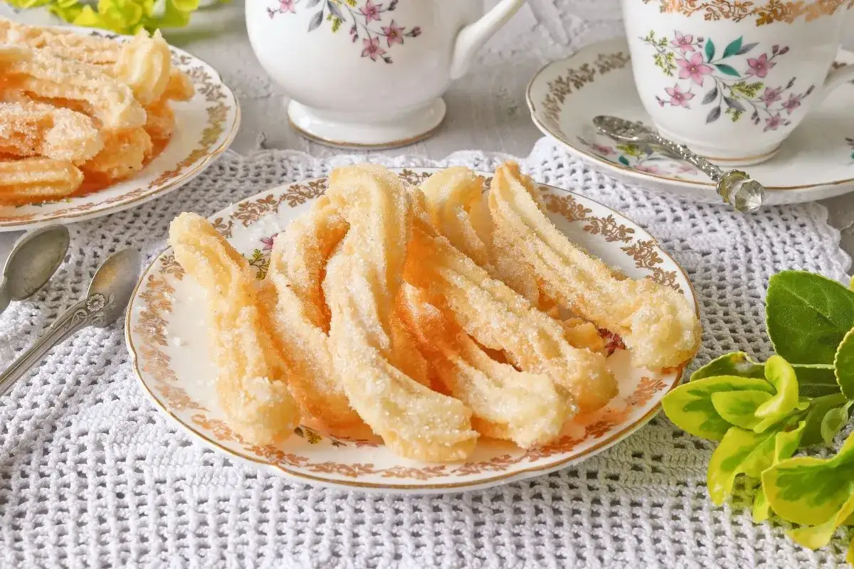 Churros sin gluten dorados y crujientes, espolvoreados con az&uacute;car, servidos en un plato con un dise&ntilde;o floral.