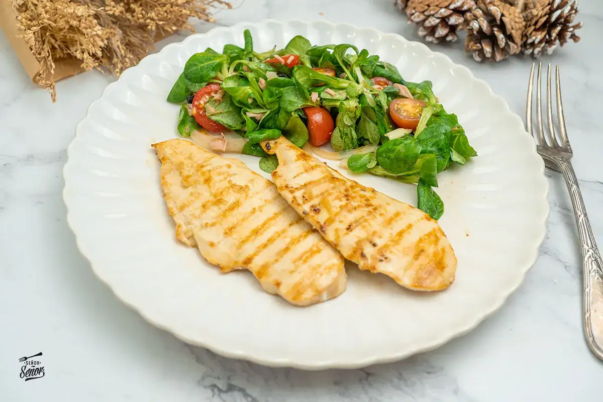 Dos pechugas de pollo a la parrilla con marcas doradas, acompañadas de una ensalada fresca con tomates y hojas verdes.
