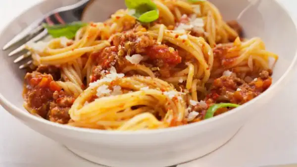 Un plato de espaguetis con carne picada y tomate, espolvoreado con queso rallado y decorado con hojas de albahaca.