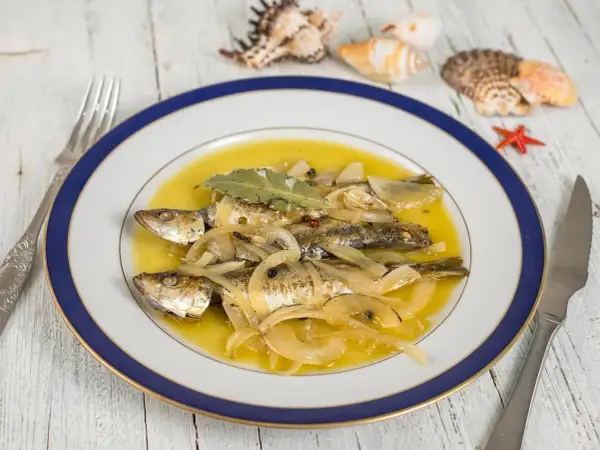 Tres sardinas en escabeche, cubiertas de cebolla y laurel, nadan en un líquido dorado. El plato está decorado con conchas marinas.