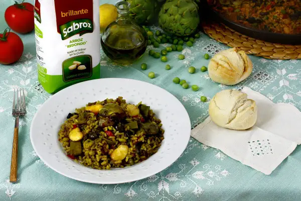 Un plato de paella de verduras, con arroz Sabroz, tomates, alcachofas, guisantes y panecillos.