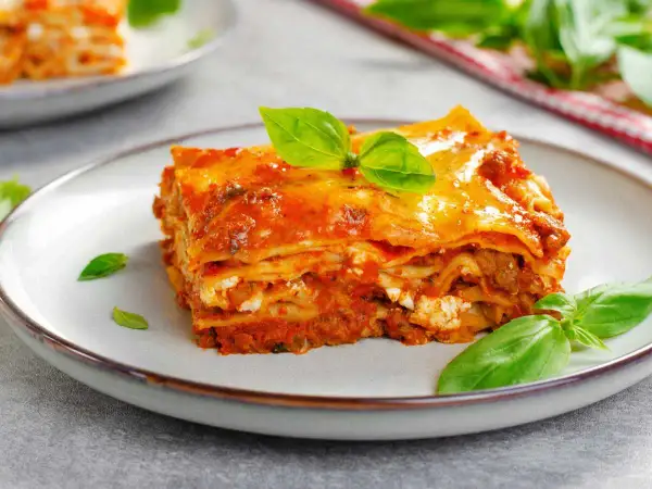 Porción de lasaña de carne con capas de pasta, salsa de tomate y queso, decorada con hojas de albahaca fresca.