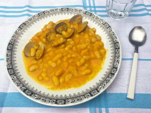 Plato de habichuelas con almejas, servido en un plato blanco con borde decorado. Acompañado de cuchara y vaso de agua.