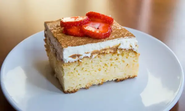 Un trozo de tarta tres leches esponjosa, cubierta con crema batida, canela y fresas frescas, servida en un plato blanco.