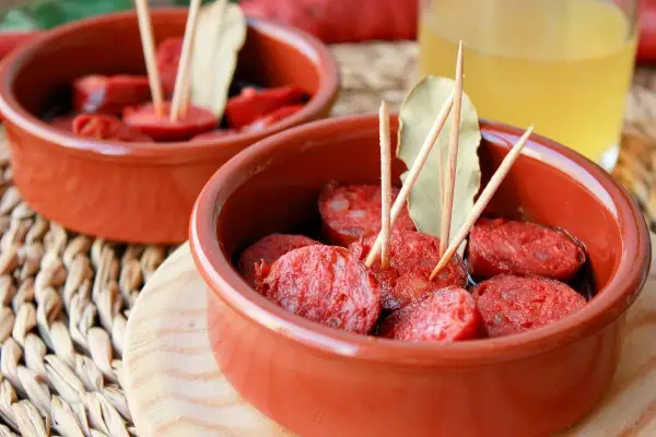 Dos cuencos de barro con rodajas de chorizo a la sidra, adornados con palillos y hojas de laurel.