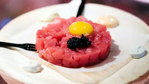Delicioso tartar de atún fresco, coronado con yema de huevo y caviar negro. Servido con salsas cremosas y un tenedor.