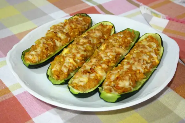 Cuatro calabacines rellenos de atún, gratinados con queso, servidos en un plato blanco sobre un mantel a cuadros.