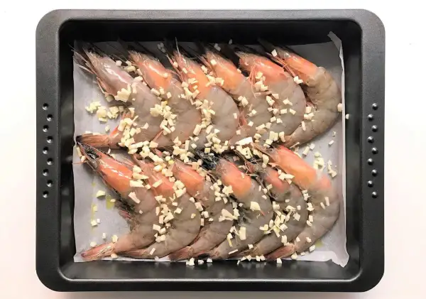 Langostinos al horno listos para cocinar, cubiertos con ajo picado en una bandeja.
