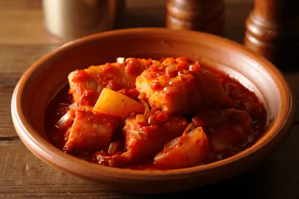 Bacalao a la vizcaina, trozos de pescado blanco en una salsa roja con pimientos y cebolla, servido en un cuenco de barro.